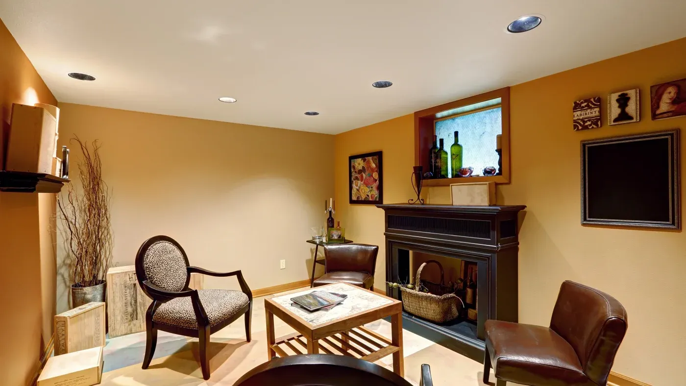Cozy living room with warm beige walls, leather chairs, and a wooden table. A fireplace is decorated with bottles and art, creating an inviting atmosphere.