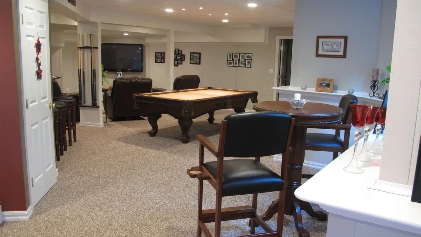 Spacious basement game room with a pool table, bar-height seating, dining table, framed photos, and warm recessed lighting.
