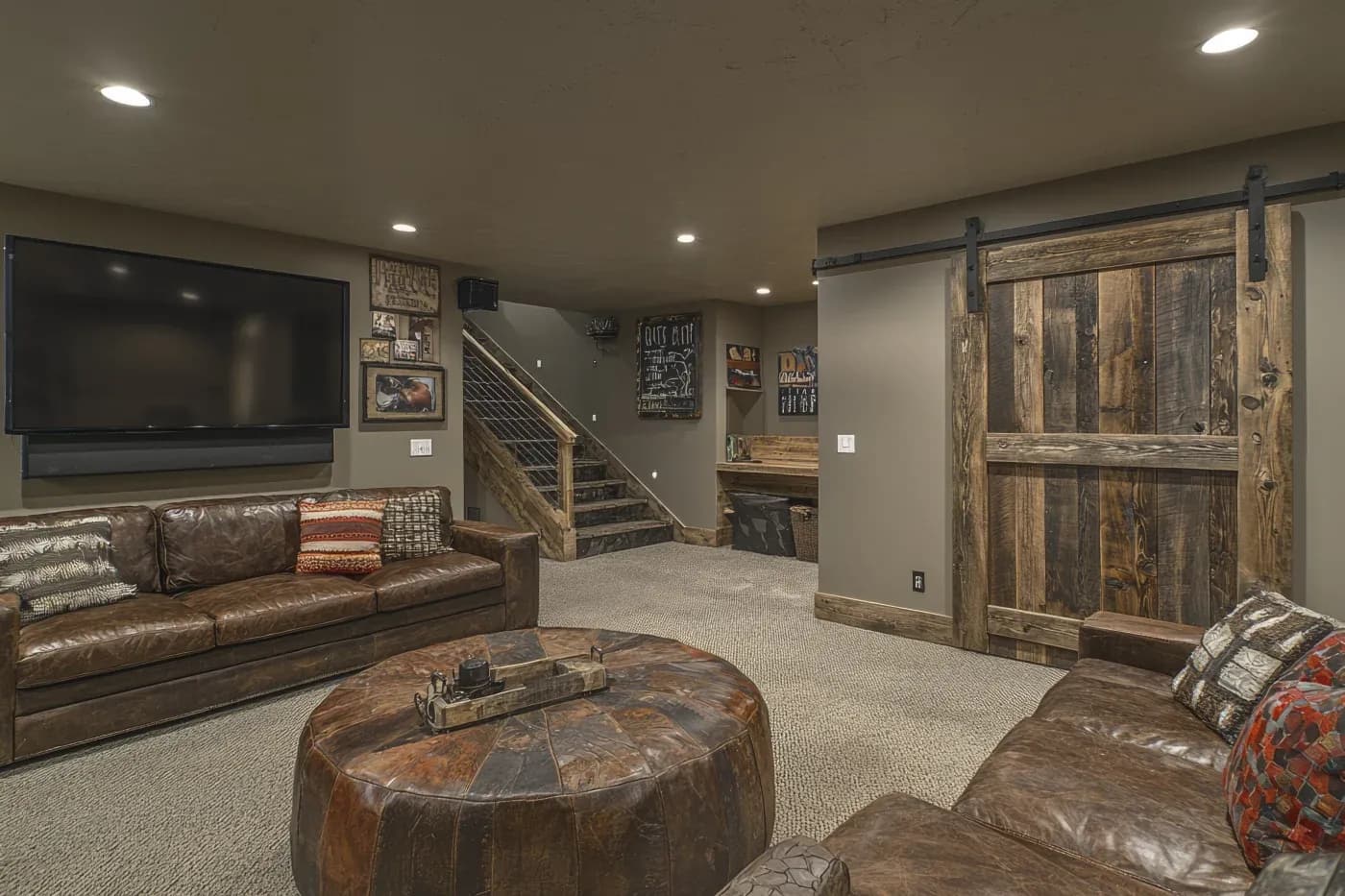 Cozy basement living room with brown leather sofas, a large TV, rustic wooden barn door, and stairs. Warm lighting creates an inviting atmosphere.