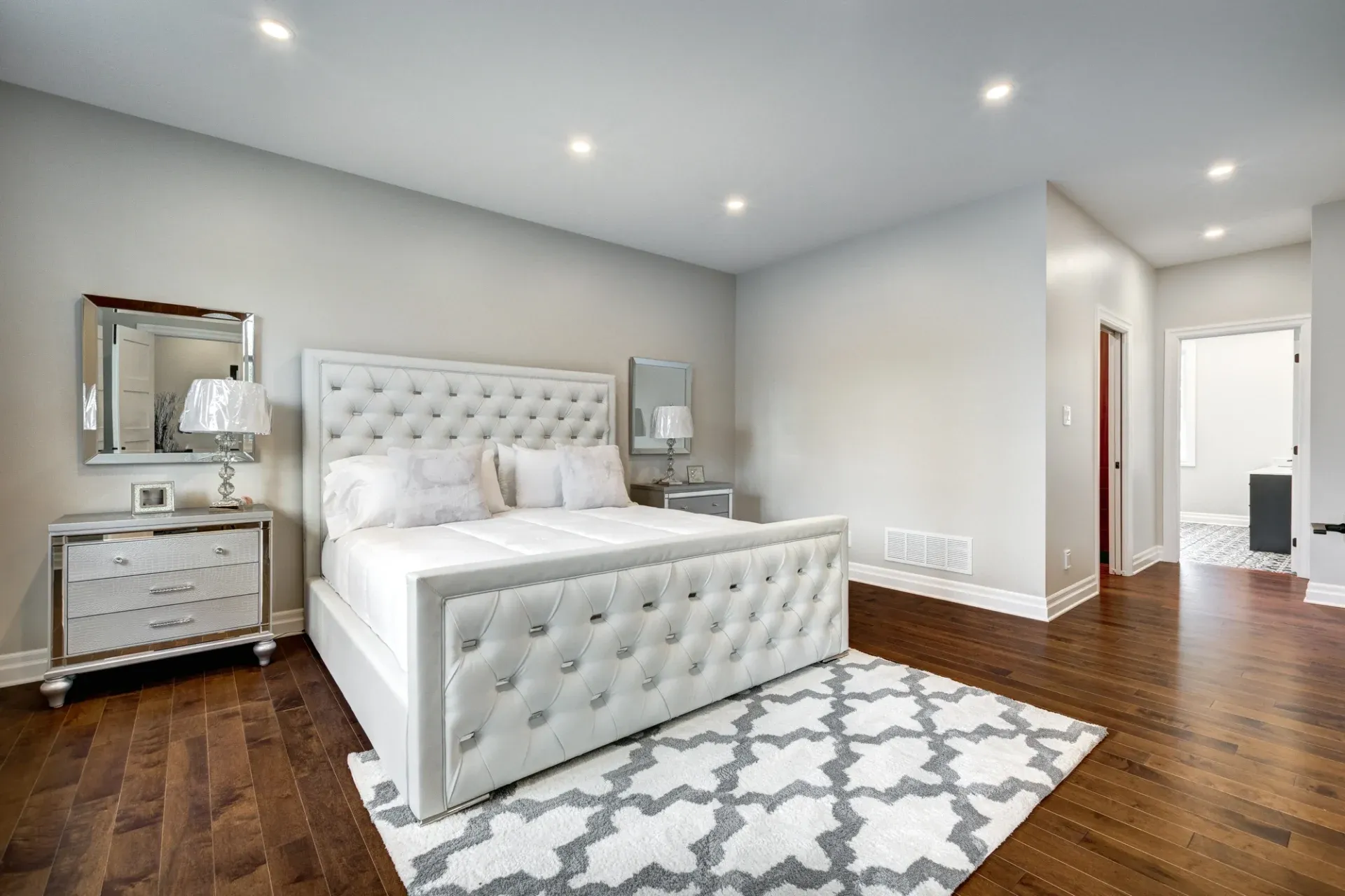 Luxurious bedroom with a plush white bed, tufted headboard, and elegant grey rug. Two mirrored nightstands add sophistication to the cozy space.