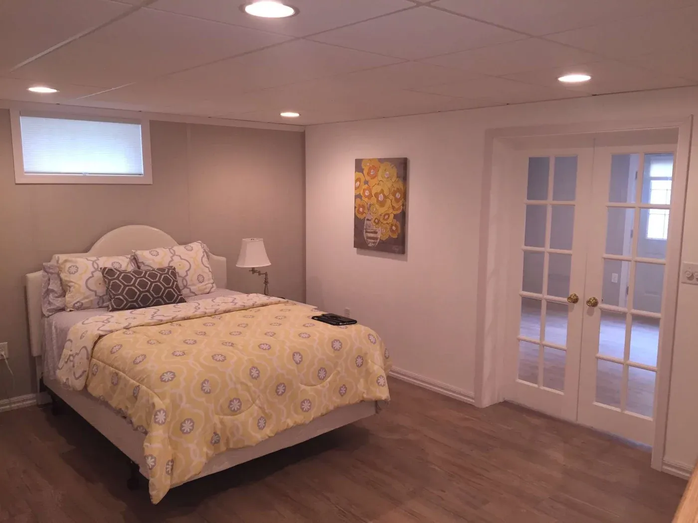 Simple bedroom with a cozy atmosphere. Features a bed with yellow floral bedding, matching artwork, a lamp, and French doors on wood flooring.