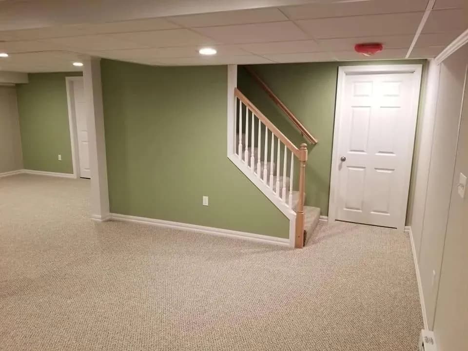 Spacious basement with beige carpet, green walls, and white paneled ceiling. Features a white door and a small staircase with wooden railings.
