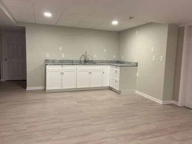 Minimalist basement kitchen with white cabinets, gray marble countertop, and light wood flooring. Soft ceiling lighting creates a clean, modern feel.