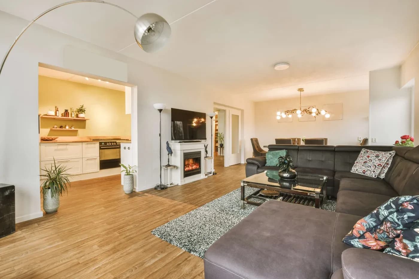Spacious living room with a modern gray sectional sofa, glass coffee table, and stylish chandelier. Open kitchen with shelving seen through an archway. Cozy, inviting ambiance.