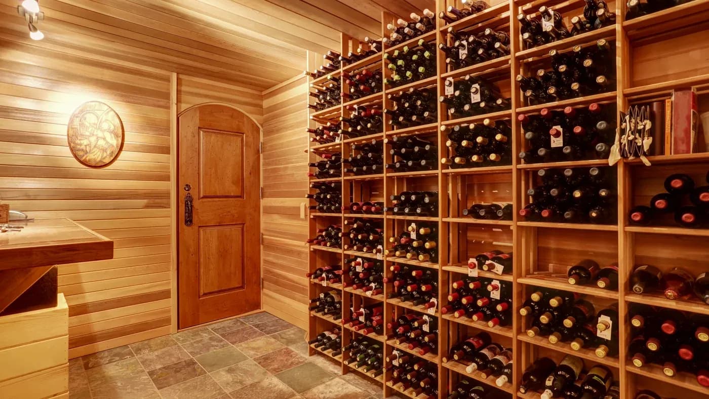Wooden wine cellar with shelves filled with wine bottles. Warm lighting, a rustic door, and a tiled floor create a cozy, elegant atmosphere.
