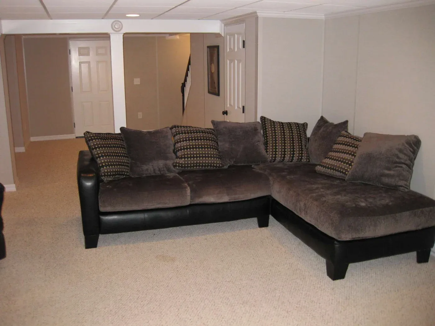 A cozy basement room with beige walls and carpet, featuring a large dark gray L-shaped sofa adorned with patterned cushions. Tranquil and inviting.