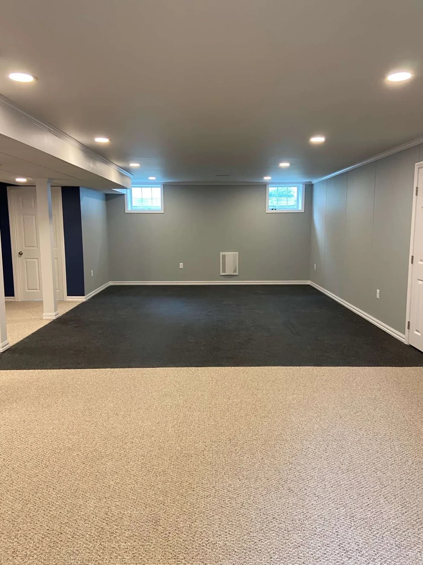 A finished basement with gray walls and recessed lighting. Two small windows let in natural light. The floor is half carpeted in beige and half in dark gray, creating a clean and modern look.