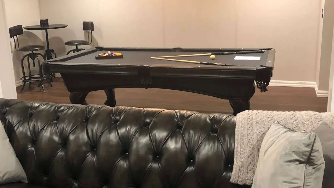 A cozy game room featuring a black leather sofa in the foreground and a pool table with arranged balls, a cue stick, and paper in the back.