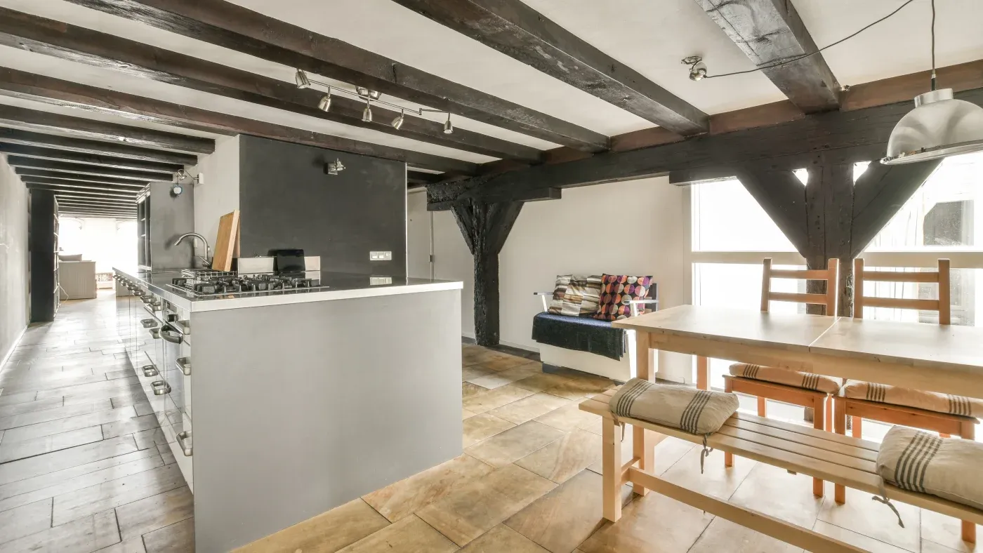 Spacious kitchen with wooden beams, light tiles, a modern gray island with a stovetop, bench seating with cushions, and a cozy sofa area.