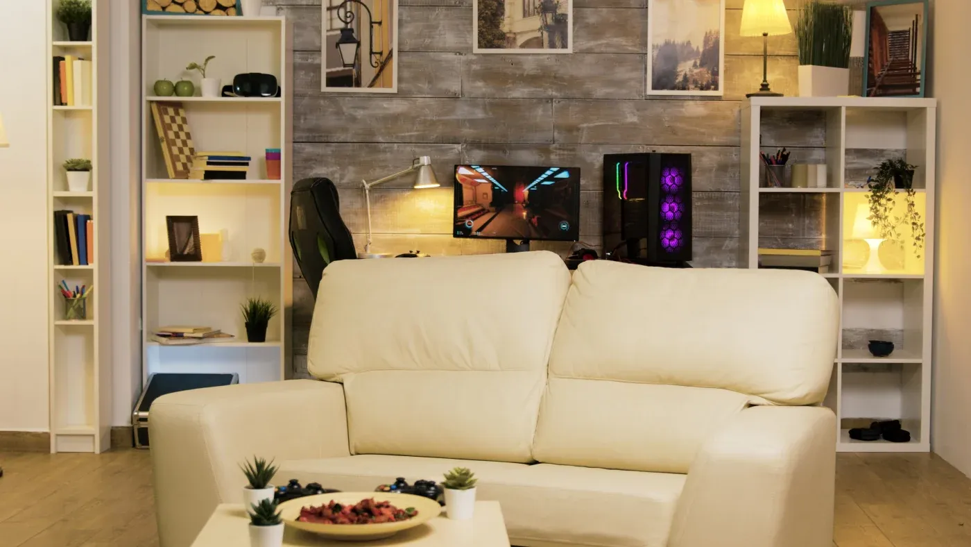 A cozy living room with a cream sofa, bookshelves, gaming setup, and ambient lighting. Decor includes plants, books, and framed art, creating a warm, stylish atmosphere.