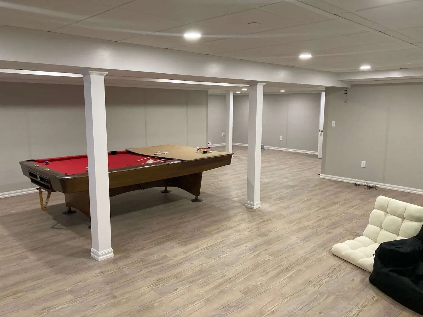 A spacious basement features light wood flooring and a red felt pool table in the center. White pillars support the ceiling, and a white cushioned chair is in the foreground. The room is well-lit with recessed lighting, creating a modern and relaxed atmosphere.